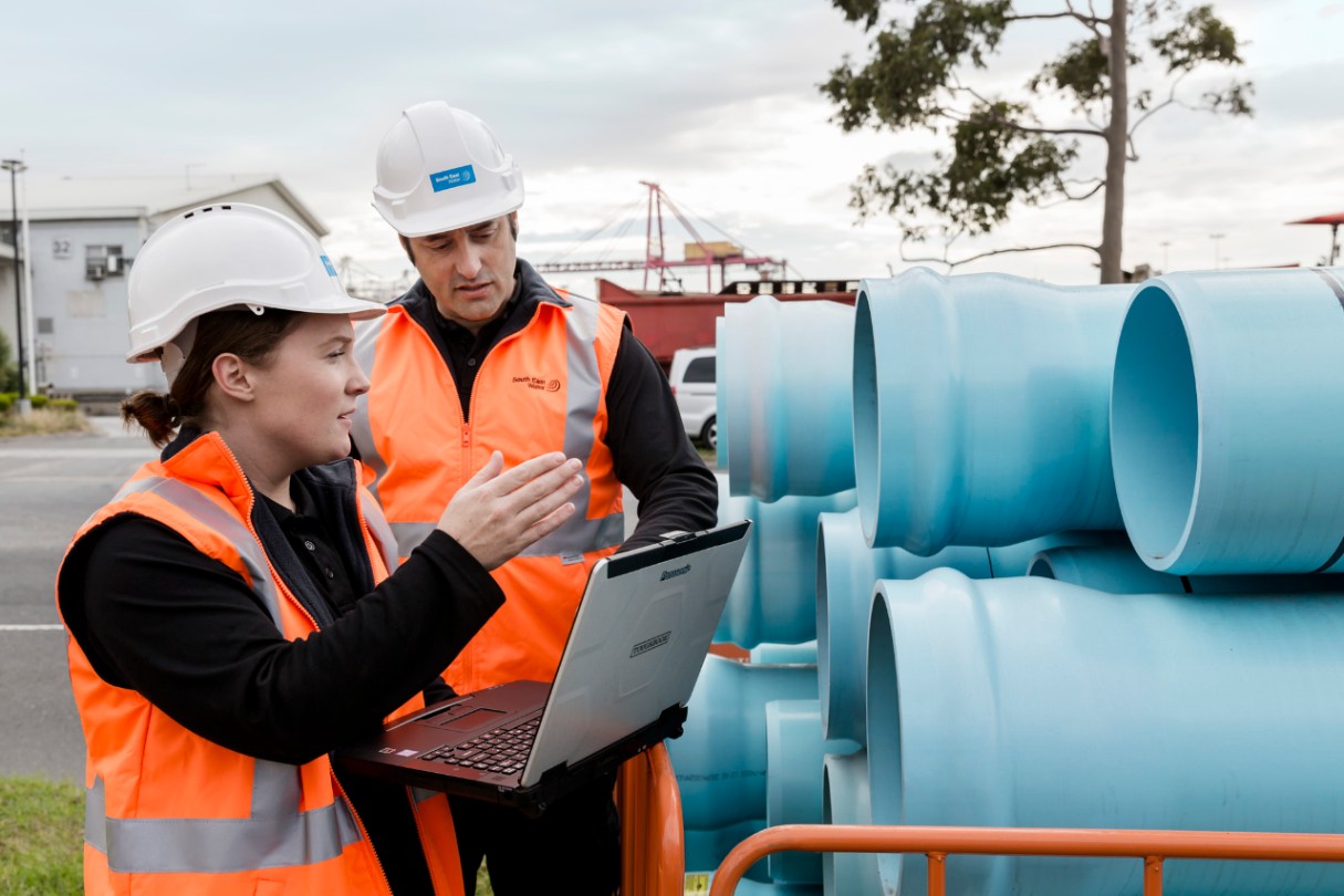 Two workers standing with a laptop looking at pipes