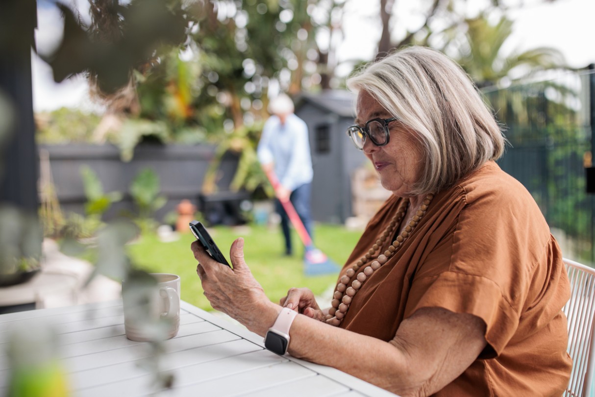 Modern elderly woman using smart phone while sitting the table in back yard, her husband is behind her cleaning the lawn.