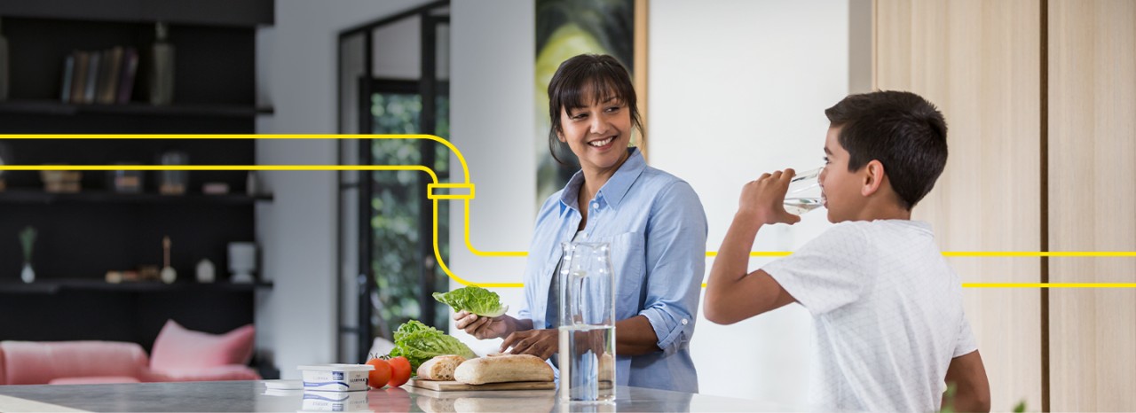 Female standing in the kitchen smiling at a boy drinking water. Photo also has a graphic of a pipe running through it. 