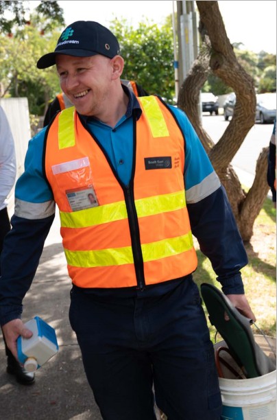 Smiling man wearing a high visibility vest carrying a water meter in one hand and a bucket in the other.  