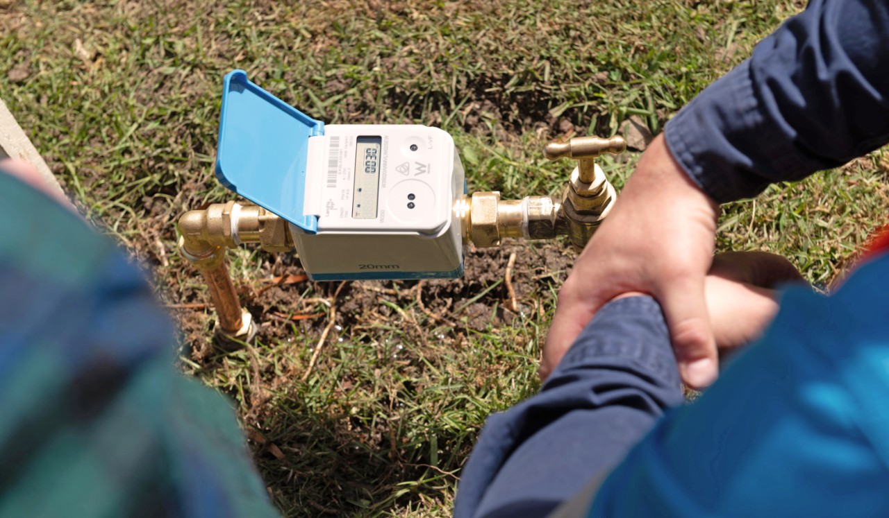 Installed digital water meter with the cover open with people standing over it. 